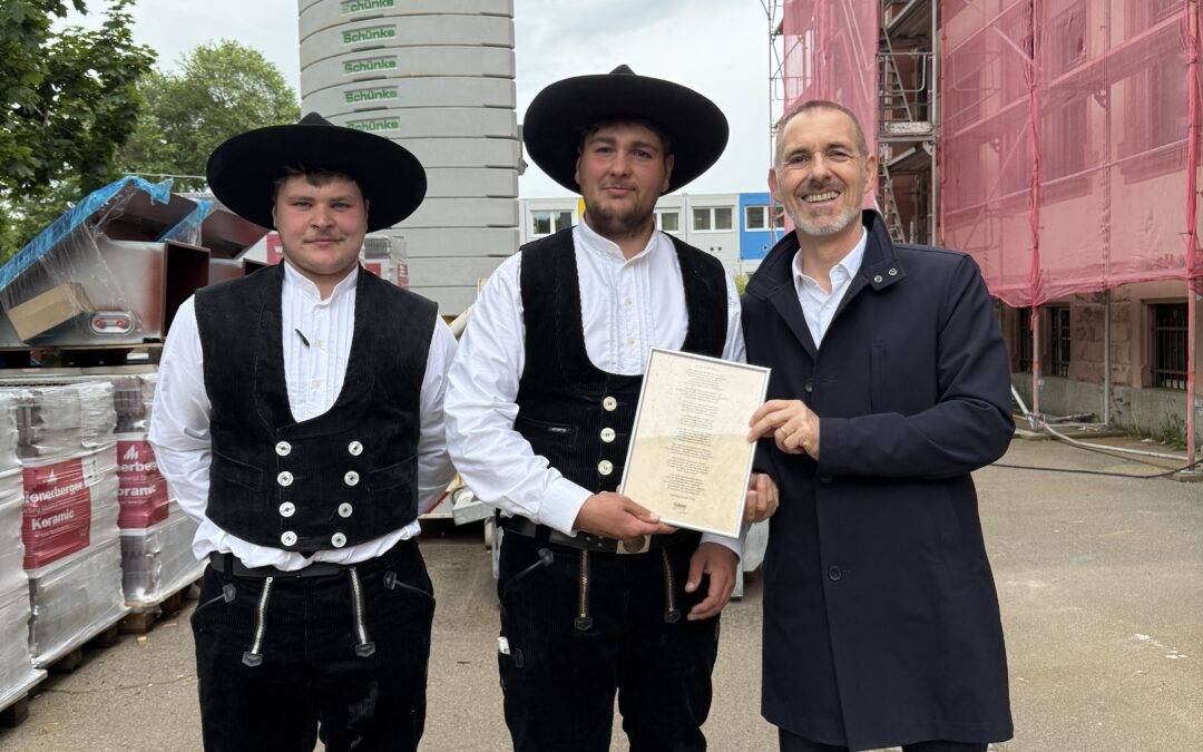 Three men posing at a construction site: two in traditional Bavarian vests and hats, and one in a dark suit holding a framed document, with pink-sheathed scaffolding and construction gear behind them.