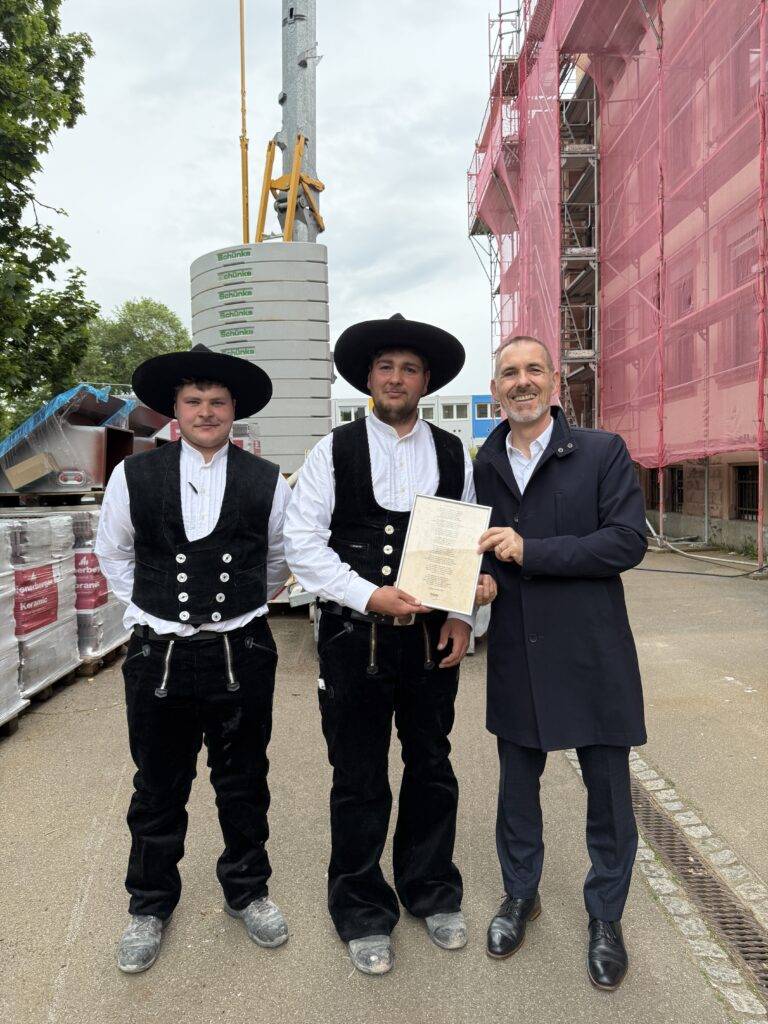 Three men posing at a construction site: two in traditional Bavarian vests and hats, and one in a dark suit holding a framed document, with pink-sheathed scaffolding and construction gear behind them.