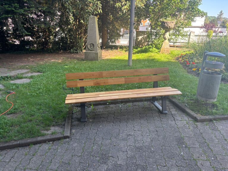 Wooden park bench on a brick path with grass, a stone monument and a decorative planter in the background, and greenery to the sides.