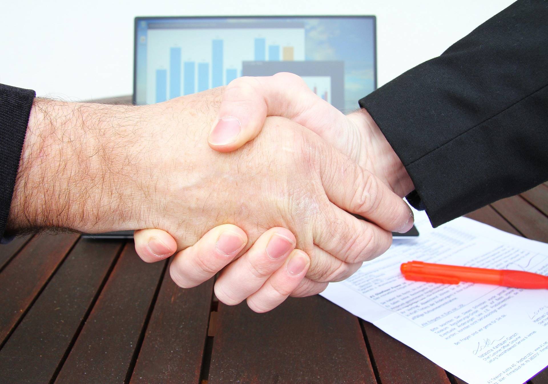 Two people shake hands over a desk with signed documents, a laptop visible in the background.
