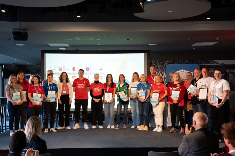 Group of people on a stage posing with certificates at an awards ceremony, with a projector screen behind them.