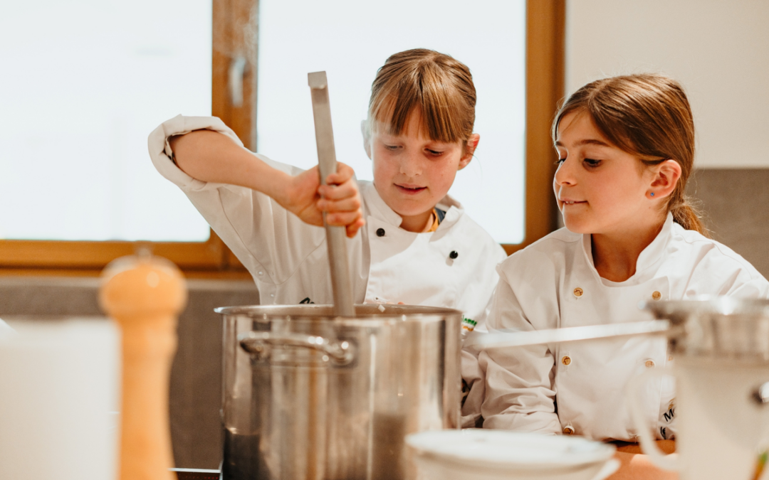 Two young girls in white chef jackets stirring a large stainless steel pot in a bright kitchen.
