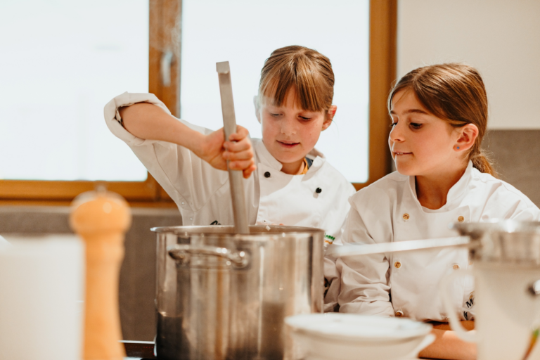 Two young girls in white chef jackets stirring a large stainless steel pot in a bright kitchen.