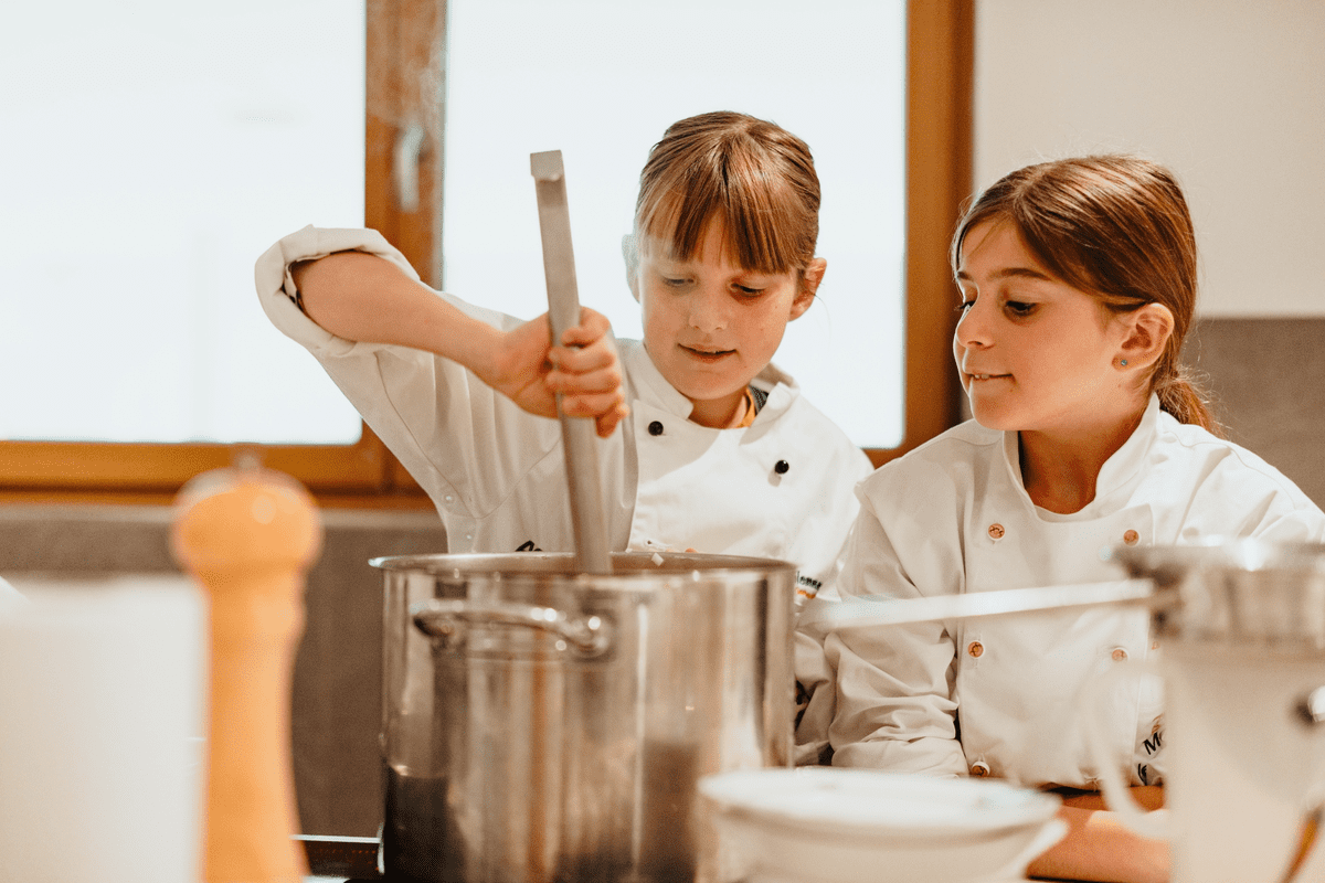 Two young girls in white chef jackets stirring a large stainless steel pot in a bright kitchen.