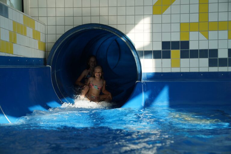 Two girls in swimsuits emerge from a blue waterslide into a pool, smiling as they slide outdoors in a water-park setting.