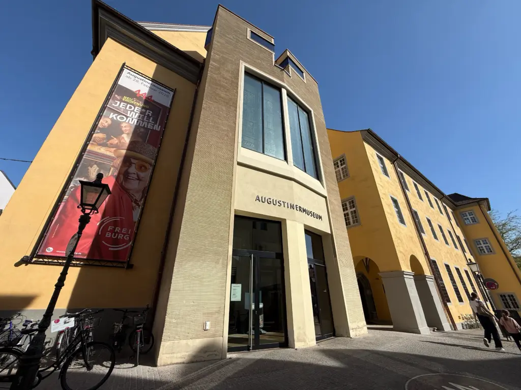 Facade of the Augustiner Museum building with beige entry, large glass windows, and a vertical exhibition banner on the left against a blue sky.
