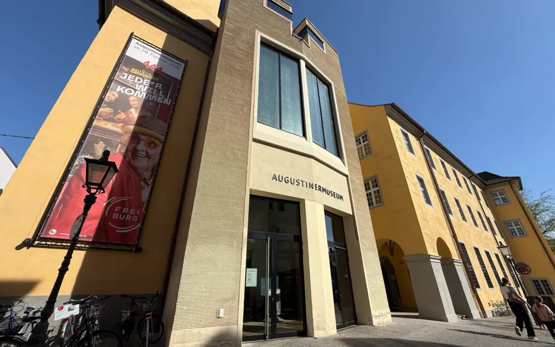 Facade of the Augustiner Museum building with beige entry, large glass windows, and a vertical exhibition banner on the left against a blue sky.