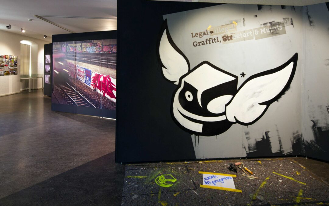 Interior of an art gallery with a large mural of a white winged cartoon shape on a dark wall, adjacent to a long photo panel of graffiti-covered train cars.