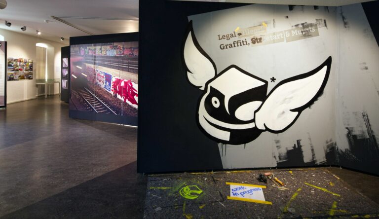 Interior of an art gallery with a large mural of a white winged cartoon shape on a dark wall, adjacent to a long photo panel of graffiti-covered train cars.