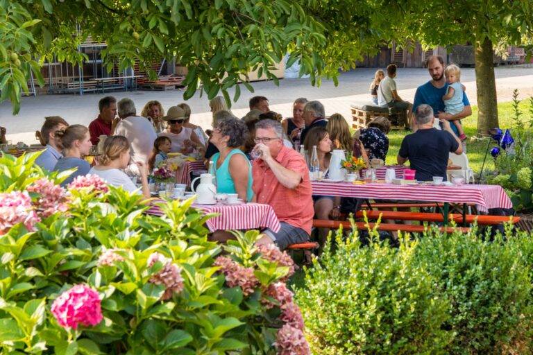 Group of people eating at long picnic tables with red-and-white striped tablecloths under a leafy tree; flowers in foreground suggest a garden party.