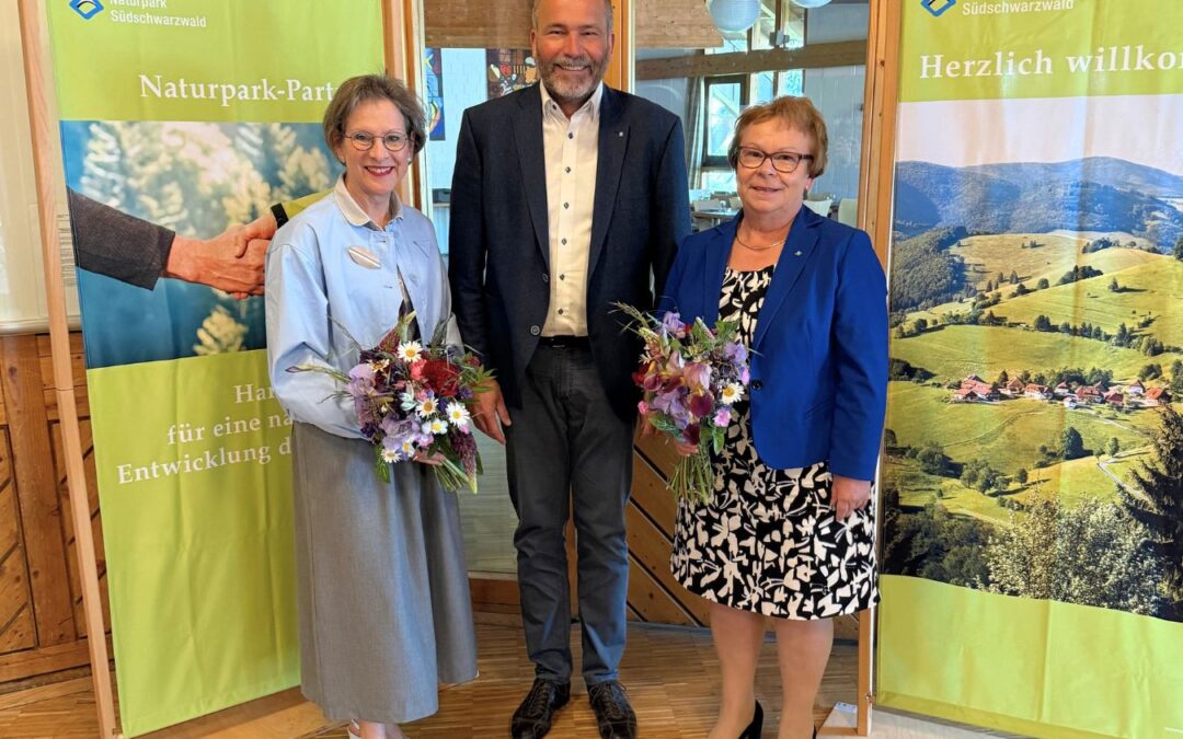 Three adults pose indoors between nature banners; two women hold colorful bouquets.
