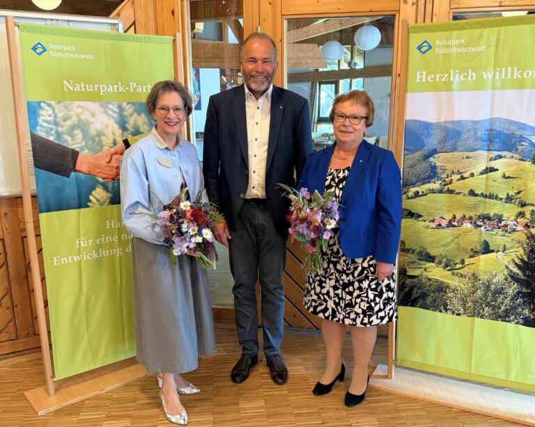 Three adults pose indoors between nature banners; two women hold colorful bouquets.