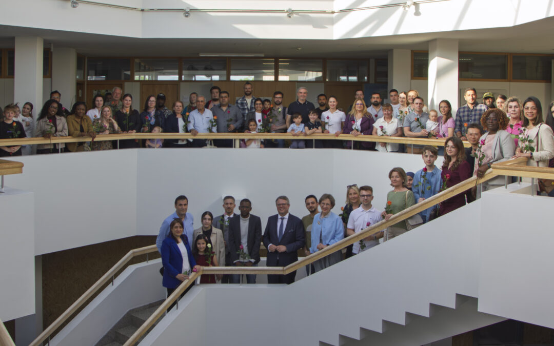 Large diverse group of adults and children posing on a modern indoor stairwell with wooden handrails, holding flowers.