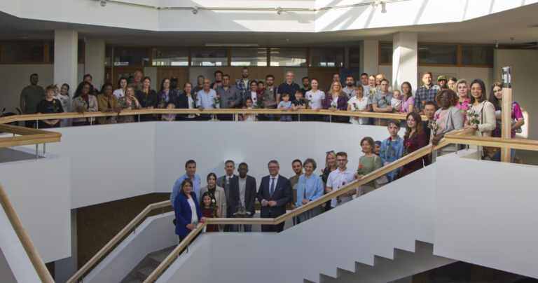 Large diverse group of adults and children posing on a modern indoor stairwell with wooden handrails, holding flowers.