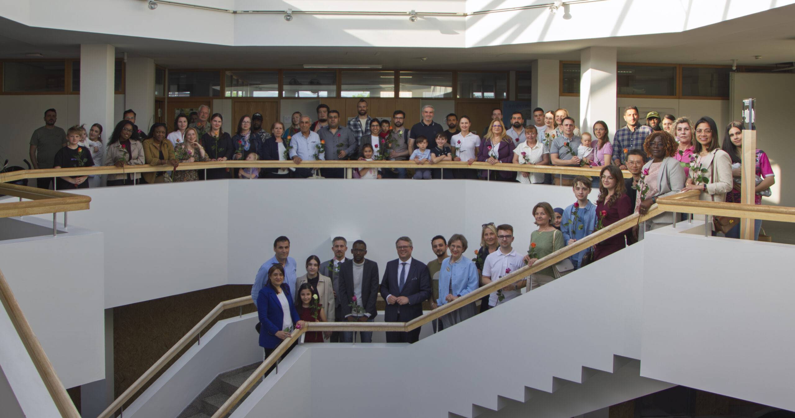 Large diverse group of adults and children posing on a modern indoor stairwell with wooden handrails, holding flowers.