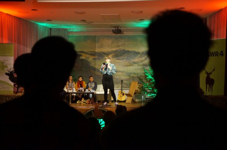 Speaker on a dimly lit stage, microphone in hand, with an audience silhouette in the foreground and a mountainous backdrop.