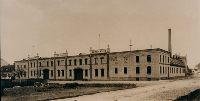 Historic brick factory building with arched windows and a tall chimney; sign reads MASCHINENFABRIK on the roofline.