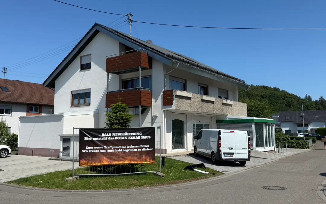 White building with brown balconies and a large banner announcing an upcoming BOTAN KEBAB HOUSE opening under a clear blue sky; a white van is parked nearby.