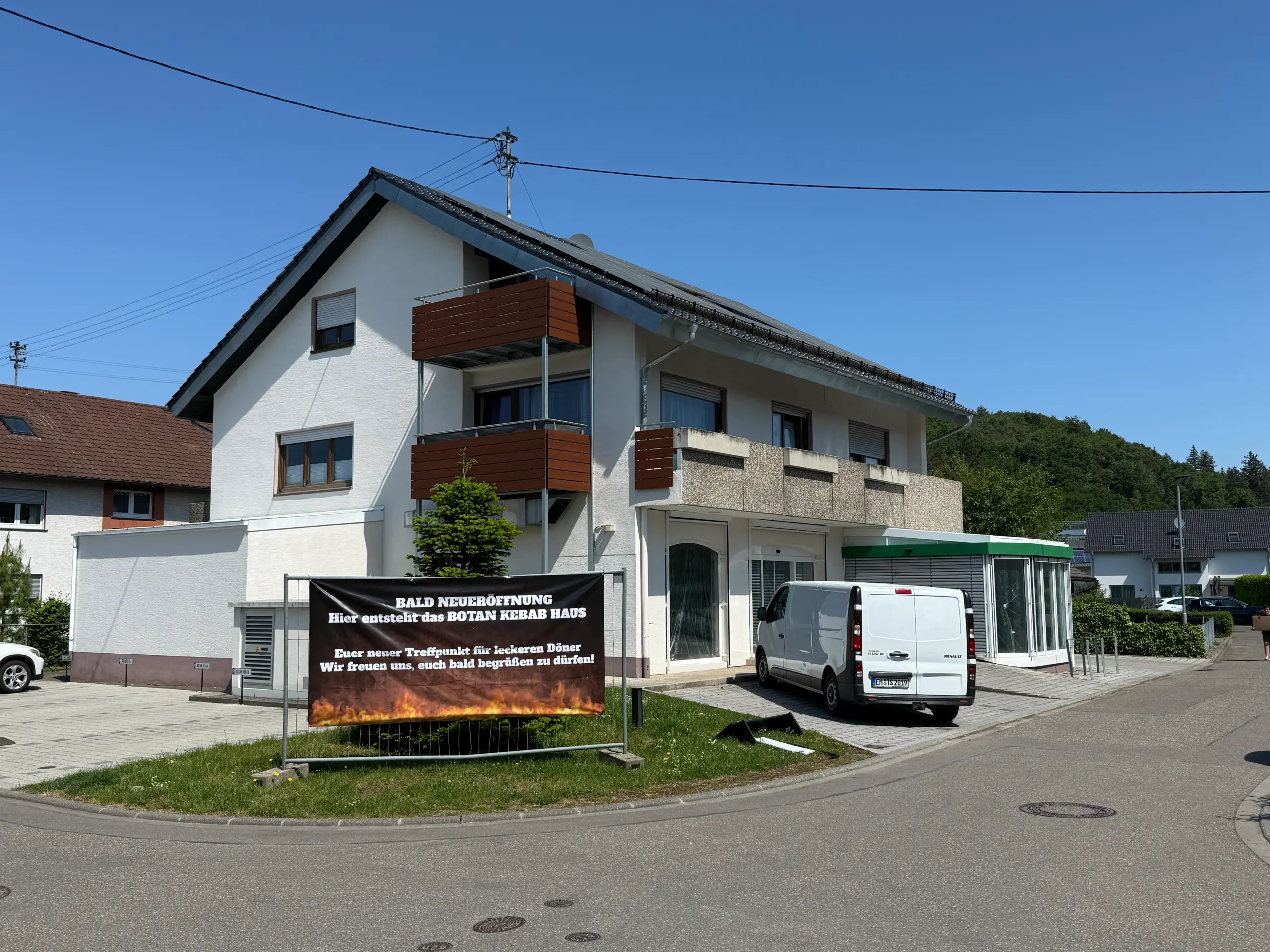 White building with brown balconies and a large banner announcing an upcoming BOTAN KEBAB HOUSE opening under a clear blue sky; a white van is parked nearby.