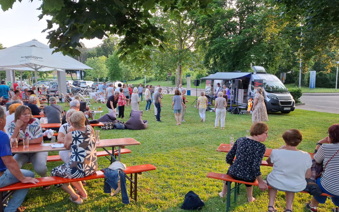 Outdoor community event in a park with people seated at red picnic tables, a white canopy, and a mobile stage/van on the right where a performer speaks or sings, crowd watching nearby.