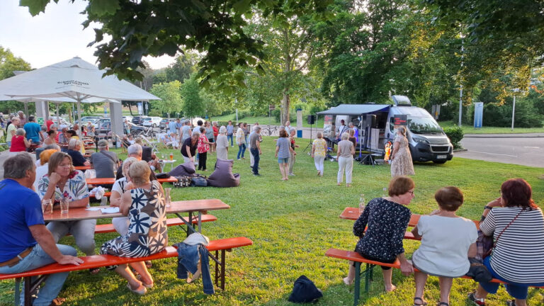 Outdoor community event in a park with people seated at red picnic tables, a white canopy, and a mobile stage/van on the right where a performer speaks or sings, crowd watching nearby.