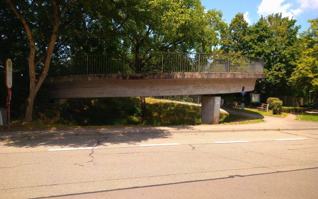 Elevated concrete pedestrian bridge with metal railing, spanning over a street lined with trees and shade beneath a blue sky.