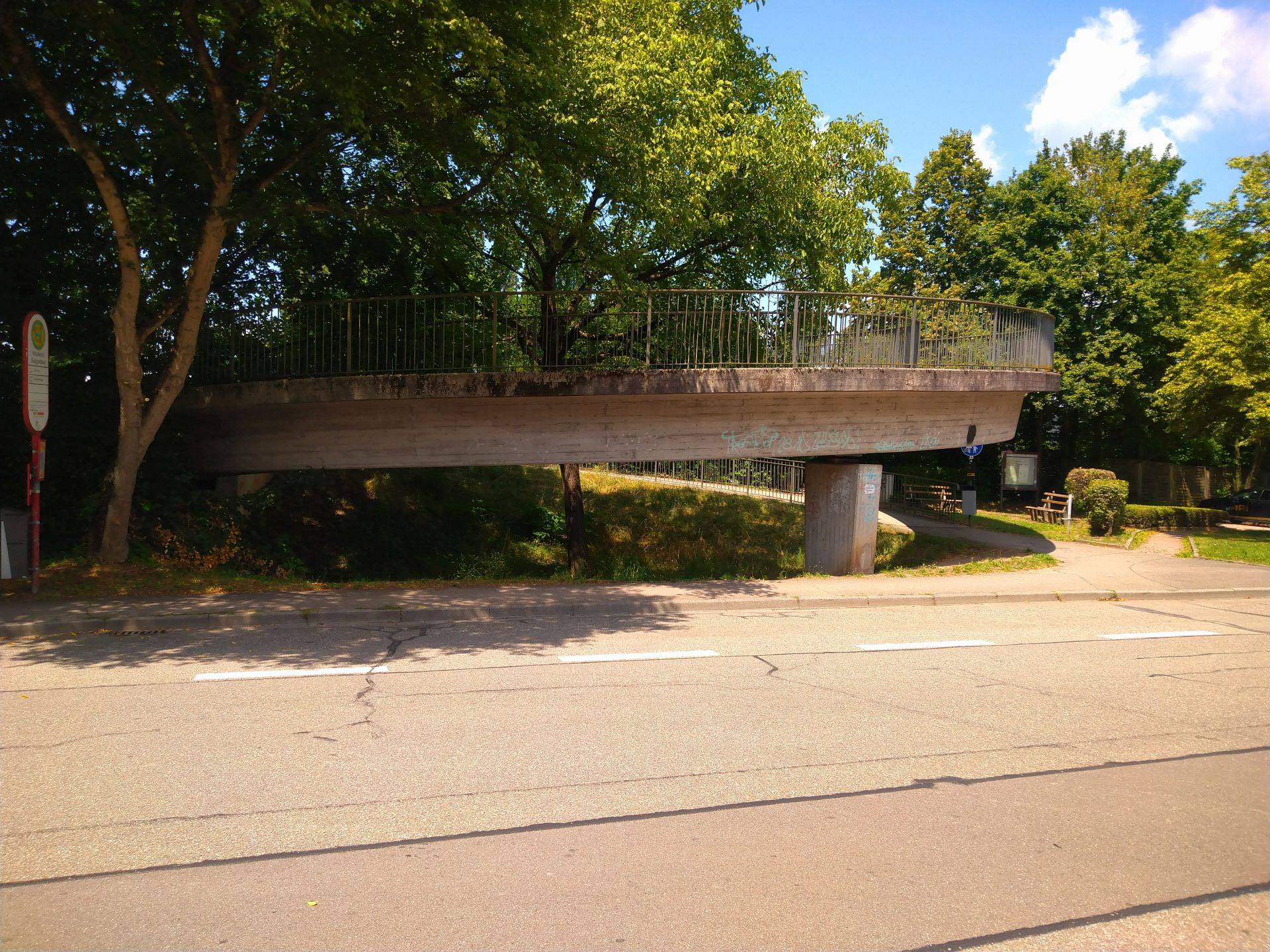 Elevated concrete pedestrian bridge with metal railing, spanning over a street lined with trees and shade beneath a blue sky.