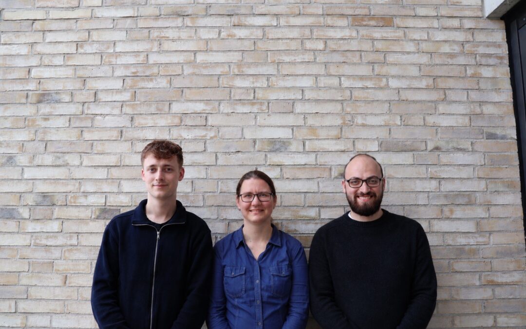 Three people posing against a light brick wall, smiling at the camera: a young man in a navy jacket, a woman in a blue shirt, and a bearded man in a black sweater with glasses.