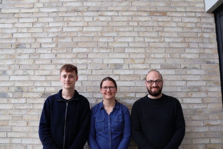 Three people posing against a light brick wall, smiling at the camera: a young man in a navy jacket, a woman in a blue shirt, and a bearded man in a black sweater with glasses.