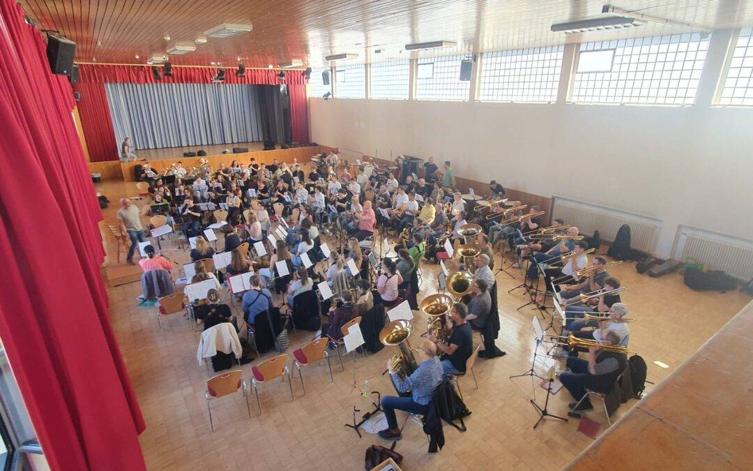 Orchestra rehearsal in a school auditorium: musicians seated with music stands, conductor at the front near a stage with red curtains.