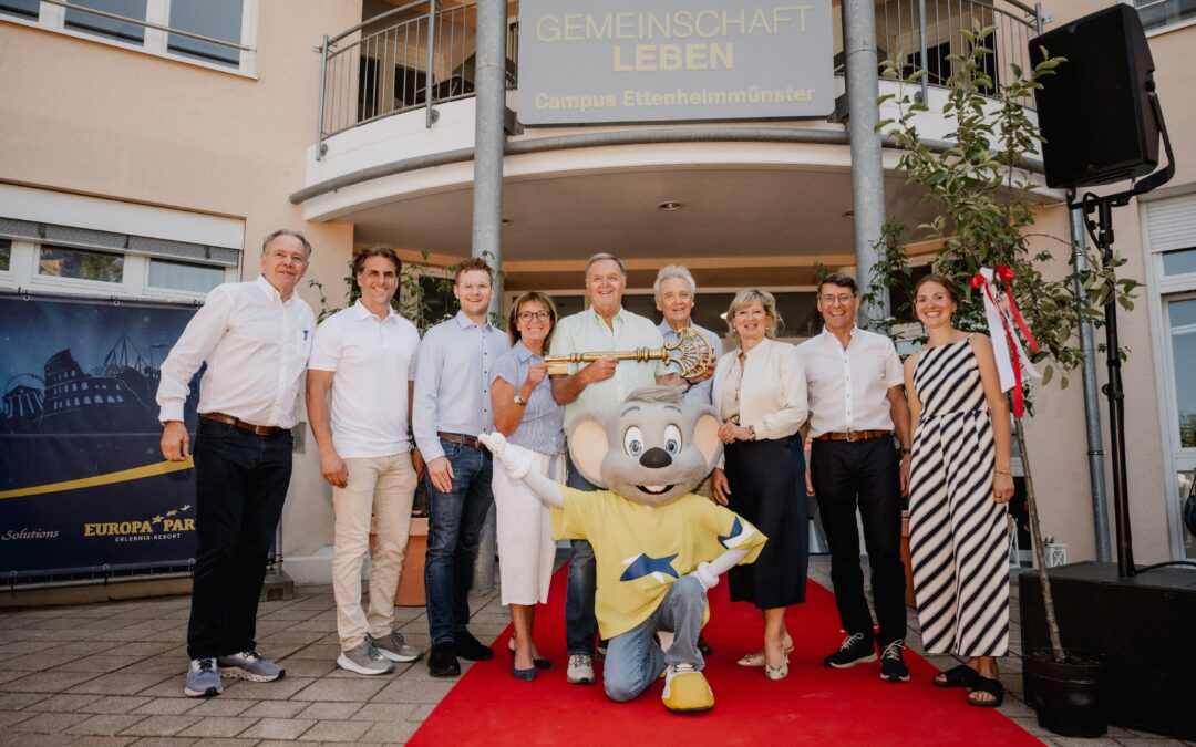 Group of adults posing on a red carpet outside a beige building with Europa Park signage and a costumed mouse character in front waving at the camera.