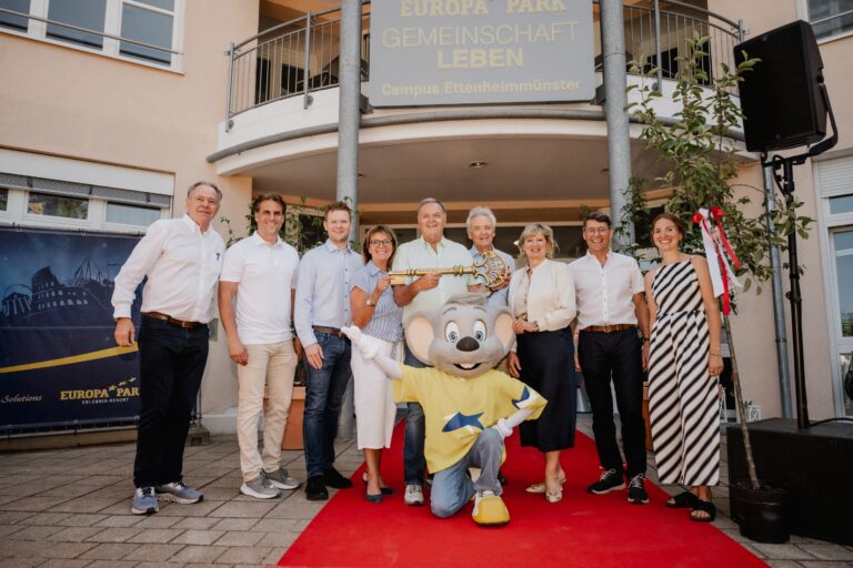 Group of adults posing on a red carpet outside a beige building with Europa Park signage and a costumed mouse character in front waving at the camera.