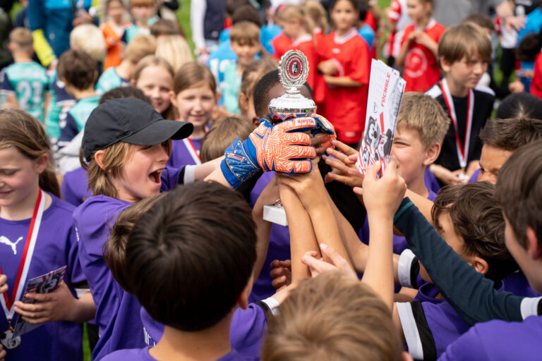 Group of children in purple shirts raise a silver trophy together amid a cheering crowd of teammates and spectators in the background.