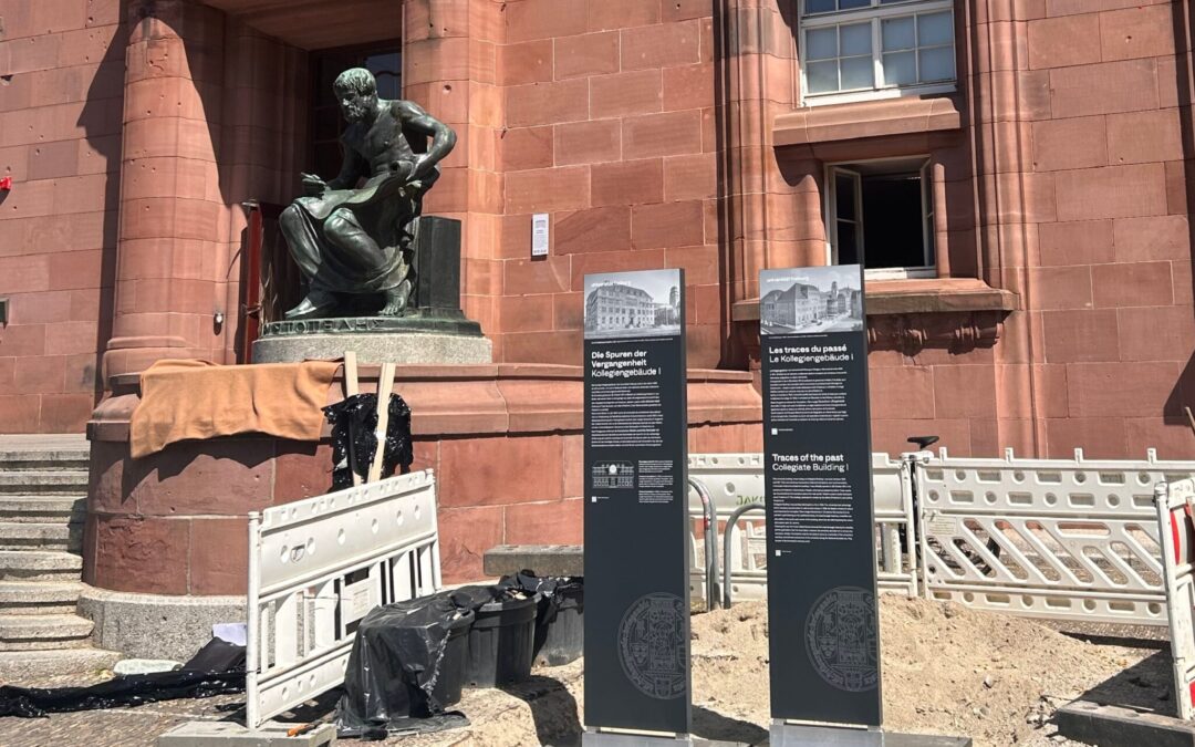 Bronze statue of a man reading a book in front of a red stone building, with construction fencing and info boards nearby