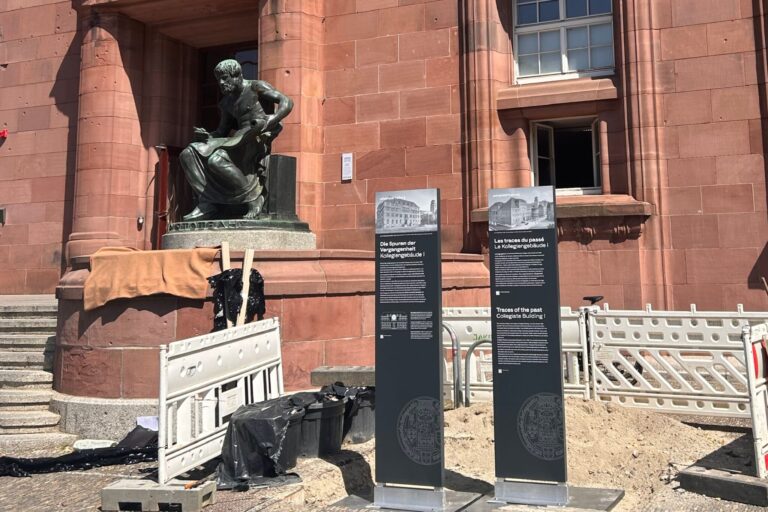 Bronze statue of a man reading a book in front of a red stone building, with construction fencing and info boards nearby