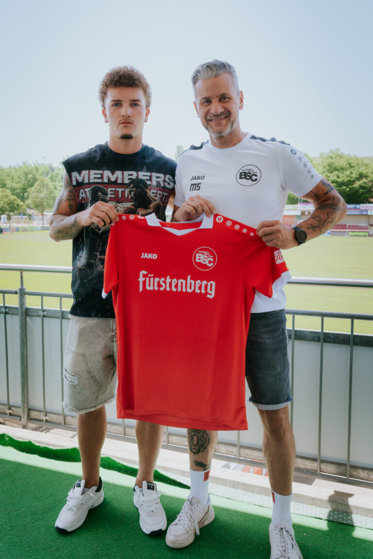 Two men stand on a railing, proudly holding a red football jersey that reads Fürstenberg, with a sports field in the background.