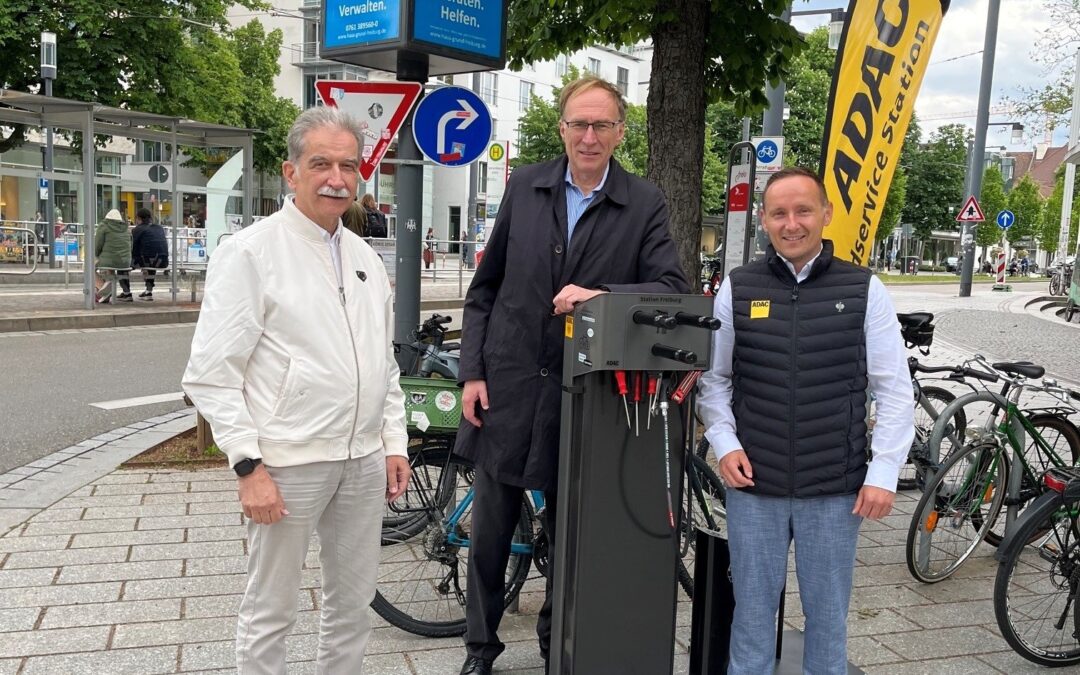 Three men stand beside a bike service/repair station on a city sidewalk, with a yellow ADAC flag nearby and bikes in the background.