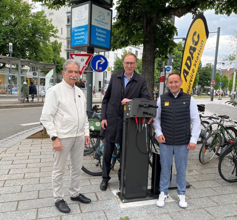 Three men stand beside a bike service/repair station on a city sidewalk, with a yellow ADAC flag nearby and bikes in the background.