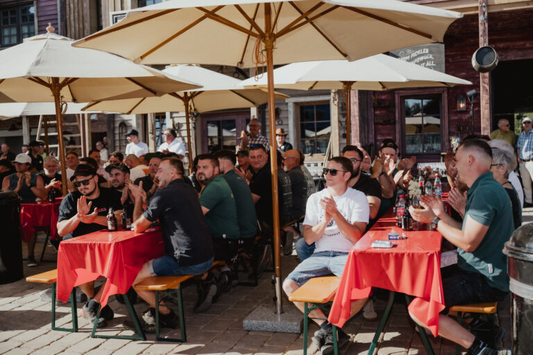 Groups of people seated at long red-covered tables outdoors, under large umbrellas, clapping during a daytime event with a rustic street backdrop.