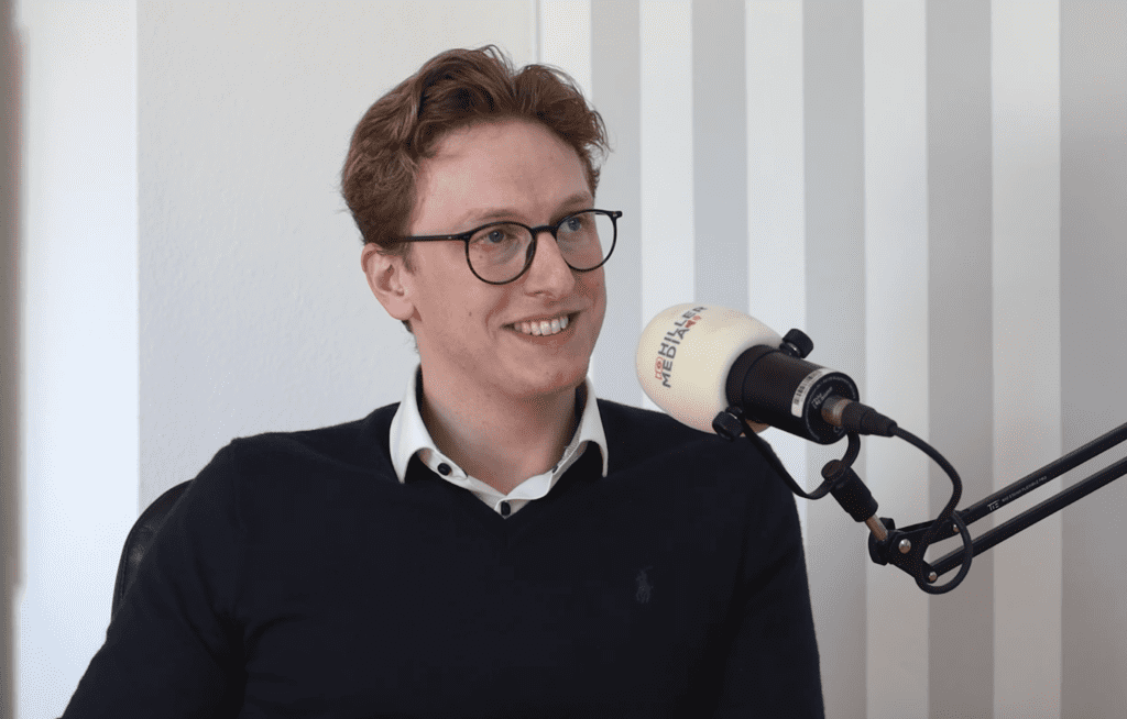 Man wearing glasses speaks into a studio microphone during a podcast interview, smiling.
