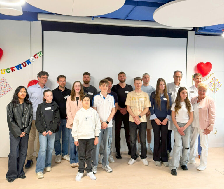 Group of students and adults posing for a photo in a decorated room with heart balloons and a colorful banner on the wall, suggesting a celebration or ceremony.