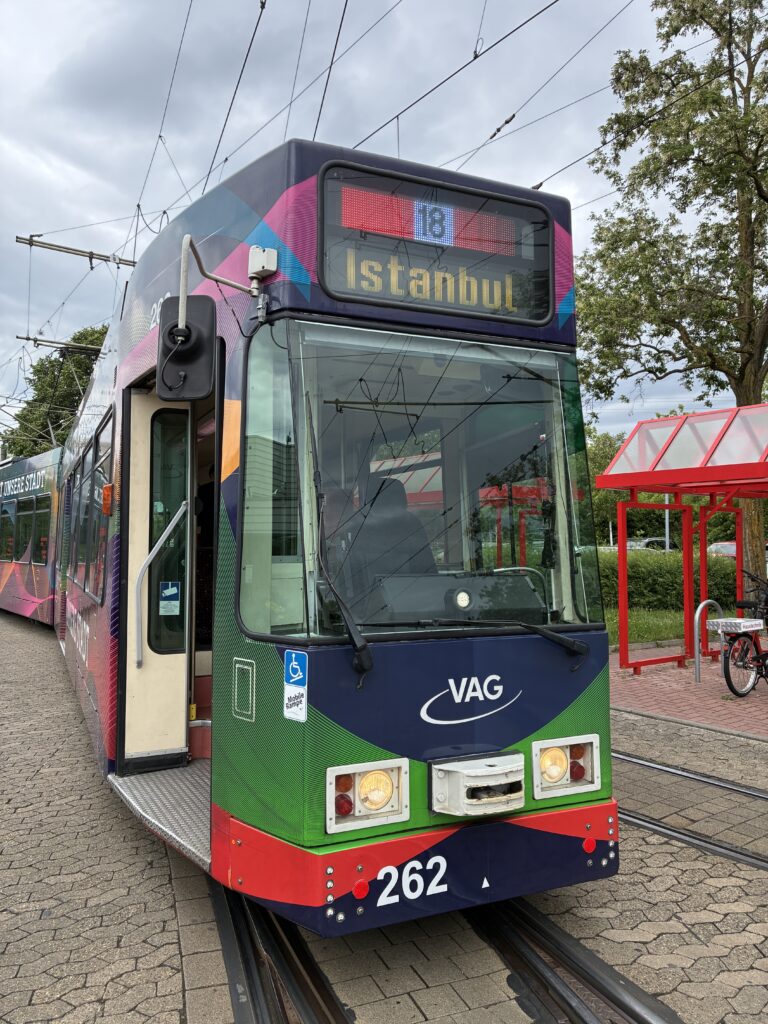 Front of a colorful tram with destination display reading ISTANBUL and route number 18, on tracks by a red shelter and trees in the background.