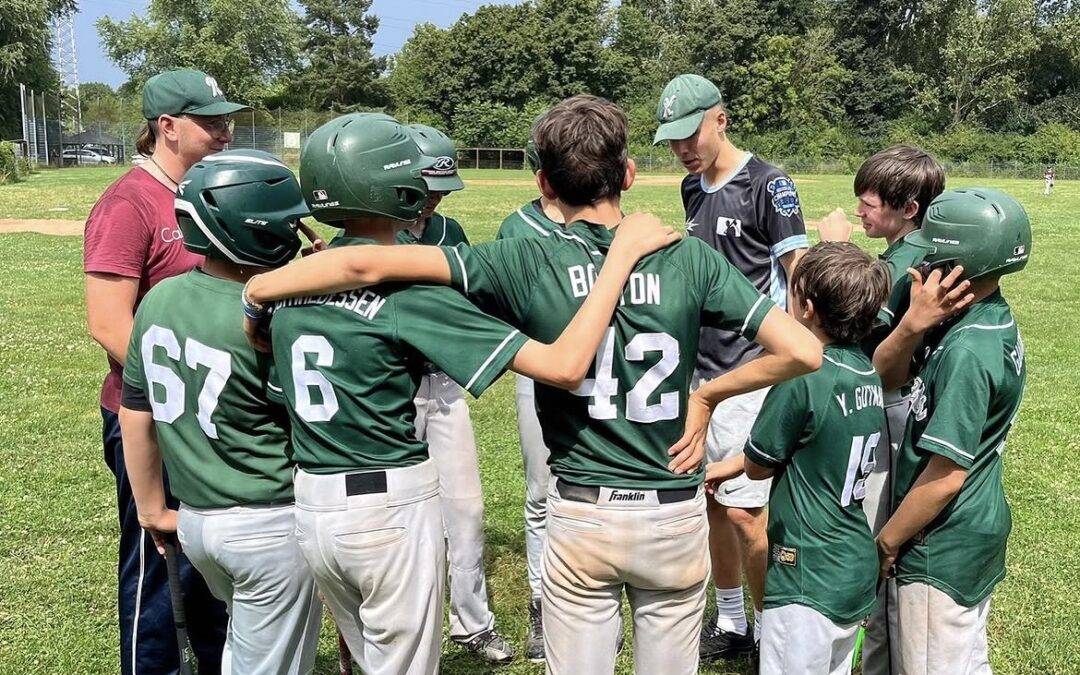 Youth baseball team in green uniforms huddled on a grassy field with helmets, wrapping arms around each other as a coach looks on nearby.