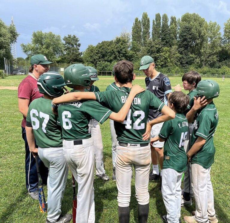 Youth baseball team in green uniforms huddled on a grassy field with helmets, wrapping arms around each other as a coach looks on nearby.
