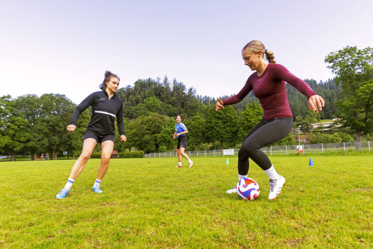 Group of young people playing soccer on a sunny park field; foreground shows a woman in a maroon long-sleeve shirt kicking a soccer ball while a teammate in black watches nearby. In the background, trees and a fence line the field.