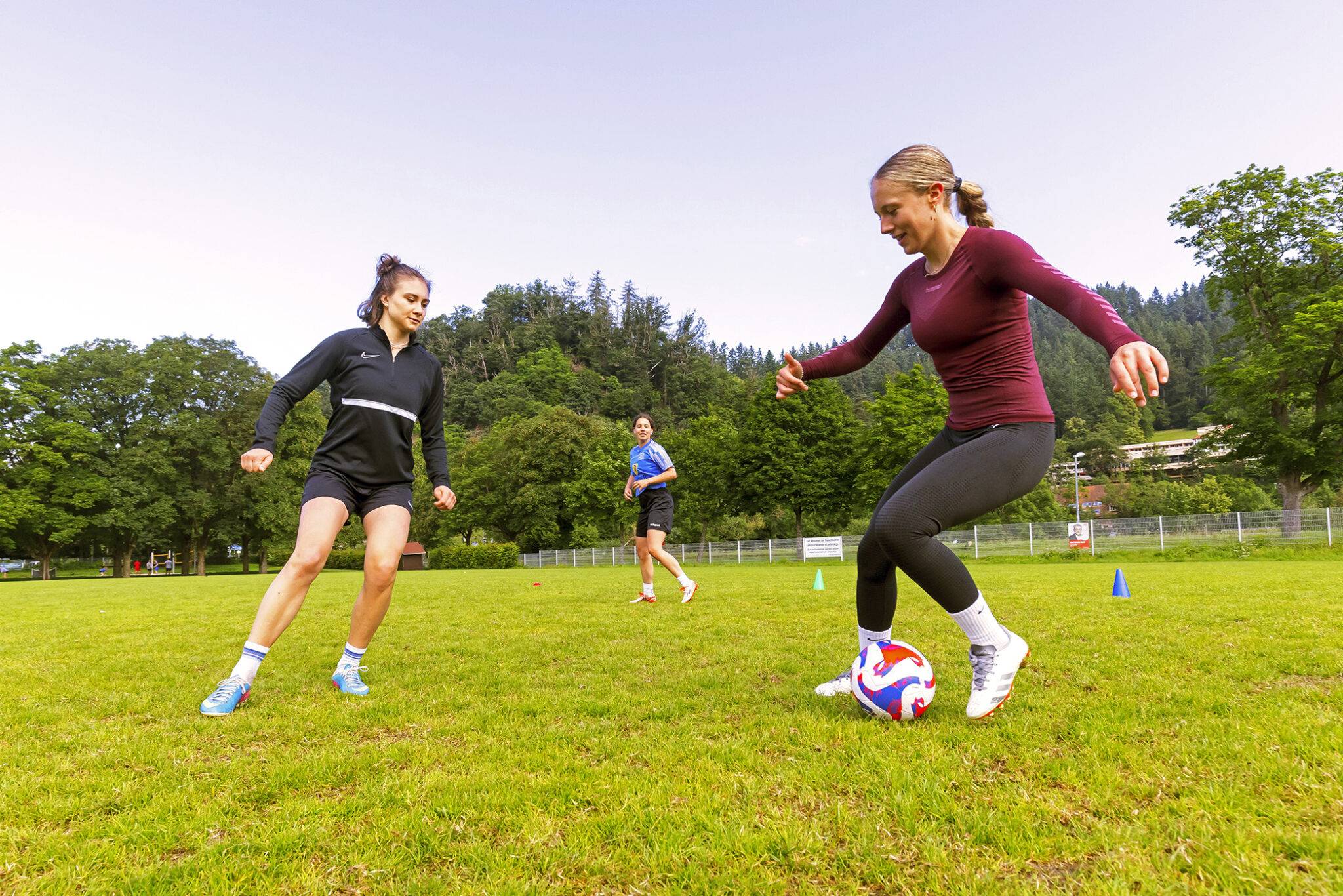 Group of young people playing soccer on a sunny park field; foreground shows a woman in a maroon long-sleeve shirt kicking a soccer ball while a teammate in black watches nearby. In the background, trees and a fence line the field.