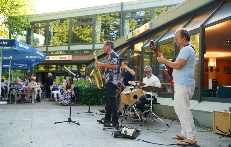 Outdoor jazz band performing in a plaza; saxophonist center, guitarist to the right, drummer behind, audience seated at tables under blue umbrellas.