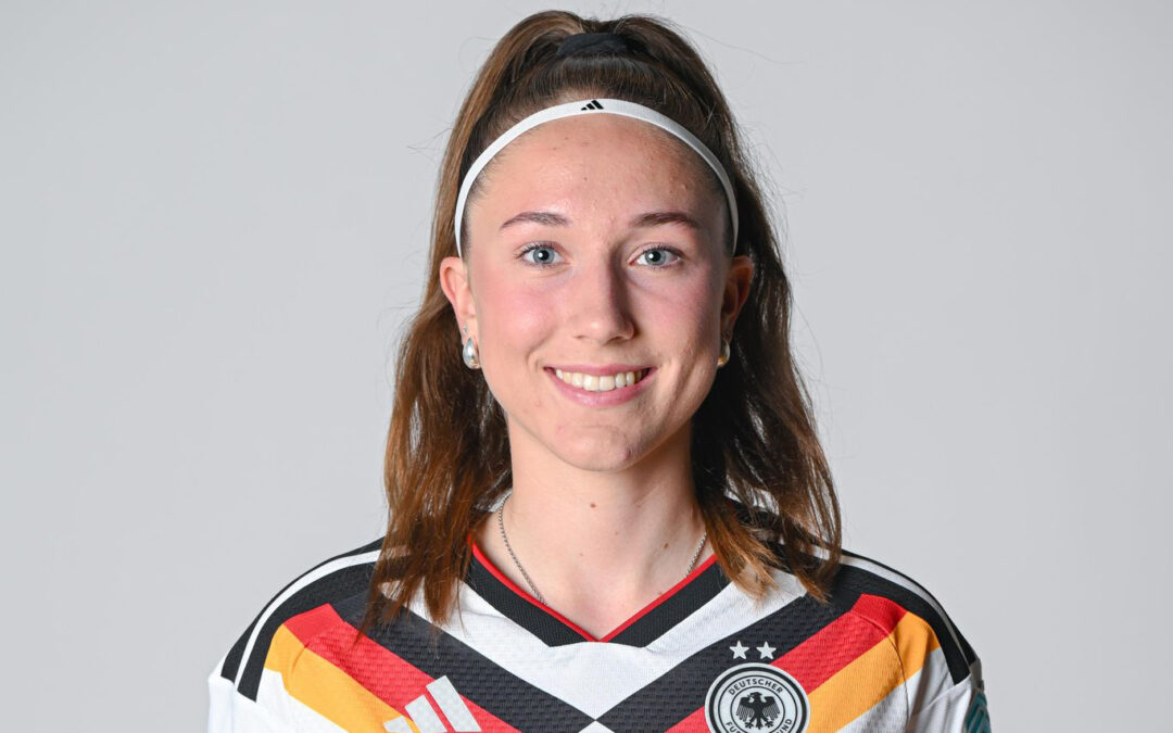 Smiling young woman with a white headband in a German national football team jersey, posing for a portrait in front of a neutral background.