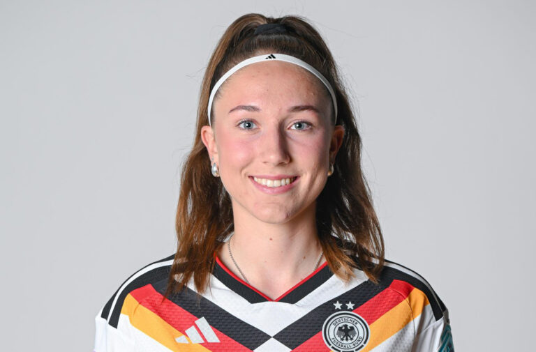Smiling young woman with a white headband in a German national football team jersey, posing for a portrait in front of a neutral background.