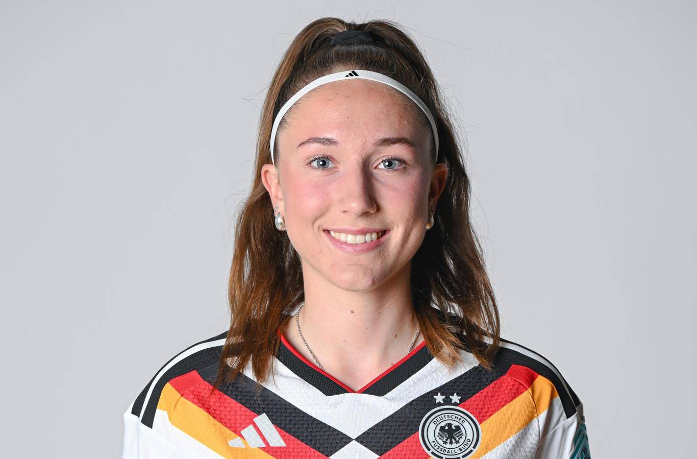 Smiling young woman with a white headband in a German national football team jersey, posing for a portrait in front of a neutral background.